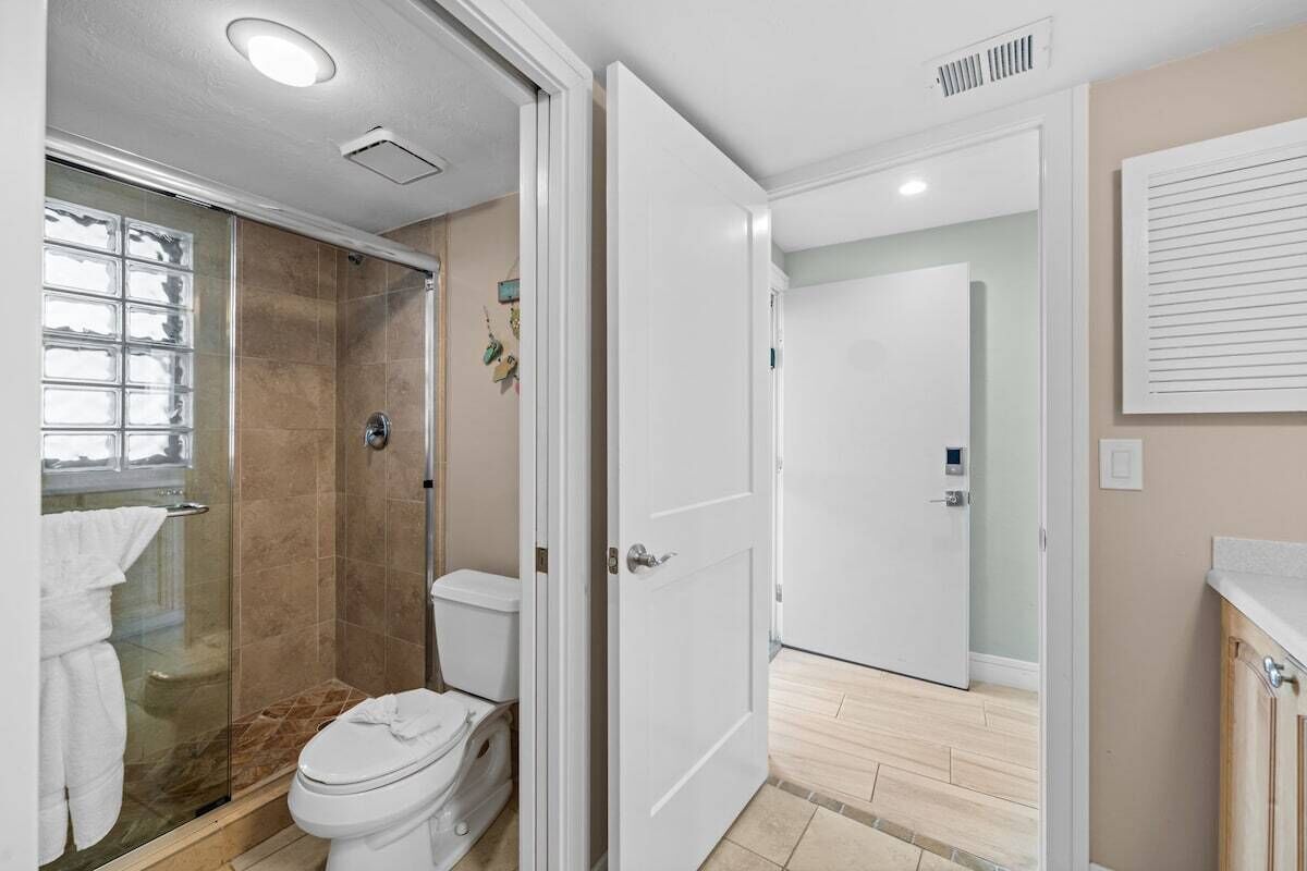 Un baño con cabina de ducha de vidrio, inodoro y puerta blanca que da a otra habitación.