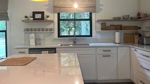 Une cuisine moderne avec des meubles blancs, un grand îlot central, un four encastré et une fenêtre donnant sur la verdure.