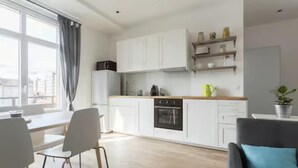 Een moderne keuken met witte kasten, een zwarte oven en een houten aanrecht. Er is een eethoek met een tafel en stoelen, en een uitzicht op de stad door grote ramen.
