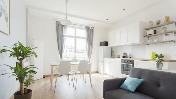 Een moderne keuken met witte kastjes, een eethoek met een tafel en stoelen, en een grijze bank met een blauw kussen.