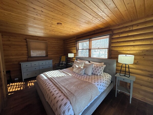 Une chambre à coucher dans une cabane en bois, avec un lit, des tables de chevet, une commode et une fenêtre avec des stores.