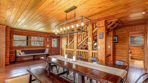 Interior de una cabaña de madera con una mesa de comedor, bancos, una escalera y un banco con vistas al exterior.