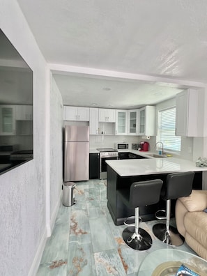 A modern kitchen with white cabinets, a stainless steel refrigerator, a microwave, and a stove. There is a small island with bar stools and a window with blinds.