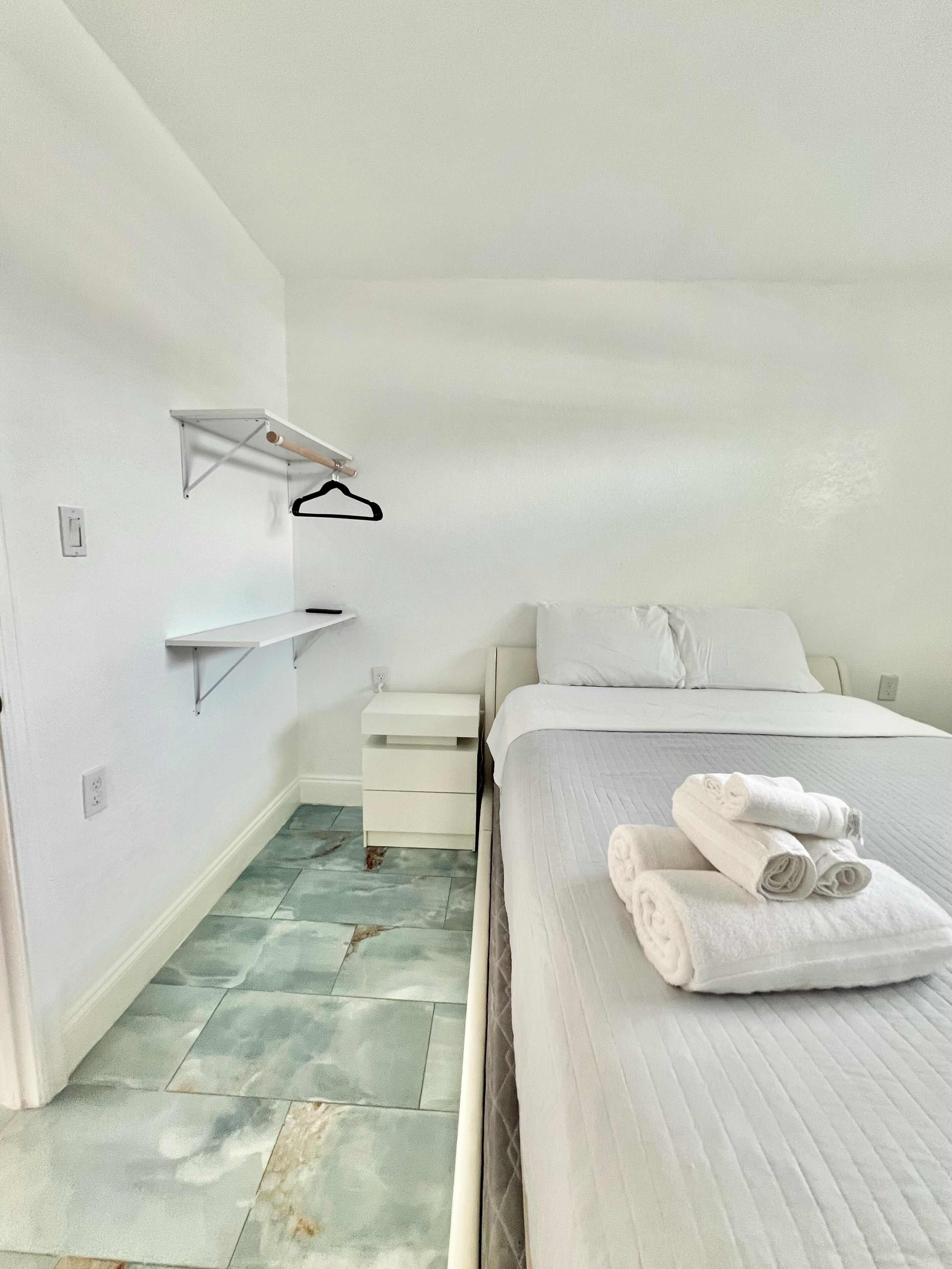 Ein schlicht eingerichtetes Schlafzimmer mit Bett, Nachttisch, Wandregal und Fliesenboden.