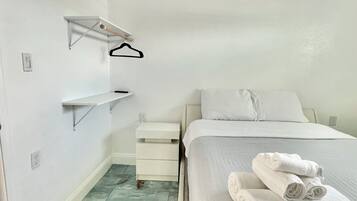 Ein schlicht eingerichtetes Schlafzimmer mit Bett, Nachttisch, Wandregal und Fliesenboden.