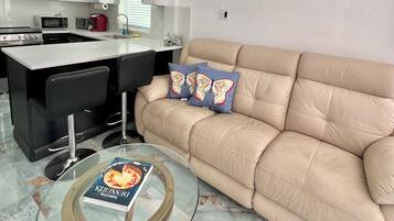 Ein modernes Wohnzimmer mit einem beige gefärbten Sofa, einem Glastisch und einer Küchenzeile im Hintergrund.