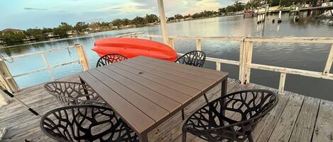 A wooden deck with a table and chairs, overlooking a calm body of water with a dock in the background.