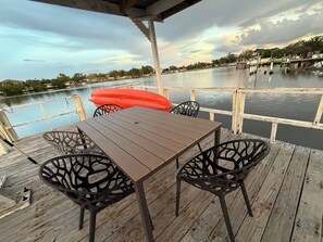 A wooden deck with a table and chairs, overlooking a calm body of water with a dock in the background. - 1bd/1ba Apartment in St. Petersburg w/PRIVATE DOCK & KAYAKS (St. Petersburg)