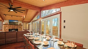 Un comedor amplio con una larga mesa de madera dispuesta para una comida, una chimenea de piedra y un ventanal con vistas a árboles.