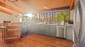 Una cocina moderna con vigas de madera en el techo, un ventanal con vistas a árboles y un refrigerador de acero inoxidable.