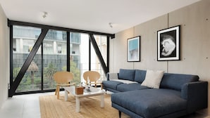 Un salon moderne avec un canapé bleu, une table basse en bois et une grande fenêtre donnant sur des bâtiments et de la verdure.