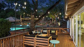 Una terraza de madera con mobiliario exterior, luces colgantes y una piscina.