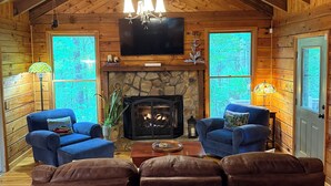 A cozy log cabin interior with a stone fireplace, two blue armchairs, a brown leather sofa, and a chandelier.