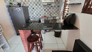 A compact kitchen with a gas stove, a sink, and a countertop. There is a refrigerator, a small stool, and a trash bin.