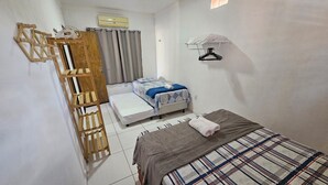 A compact room with a bed, a small table, a wooden rack, and a hanging clothes rack.
