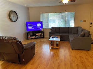 A living room with a sofa, a recliner, a coffee table, a flat-screen TV, a clock, and a window with blinds.