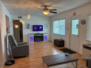 A modern living room with a flat-screen TV, a ceiling fan, a fireplace, and a large window with blinds.
