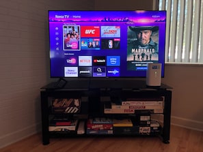 A television displaying a Roku TV interface with various streaming services and a game console on a shelf.