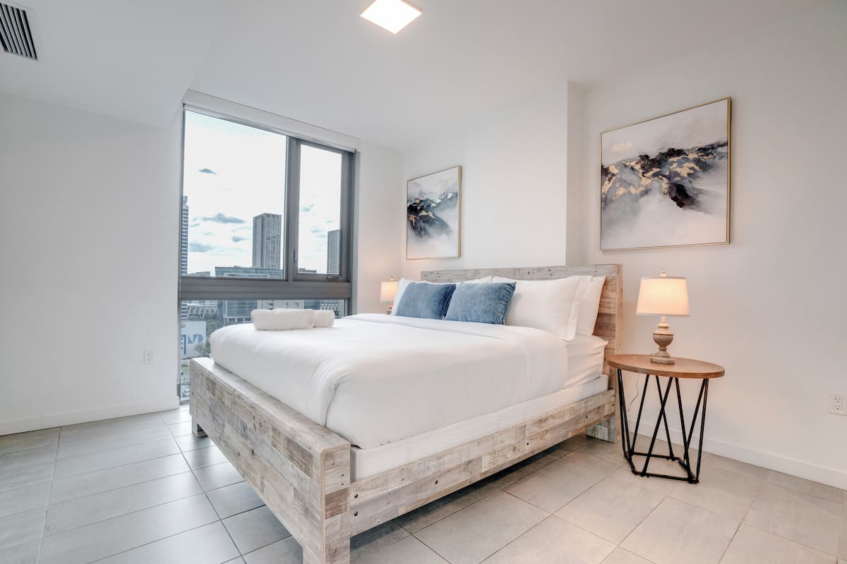 Un dormitorio moderno con una cama grande, una mesita de noche pequeña y vistas a la ciudad a trav és de un ventanal amplio.