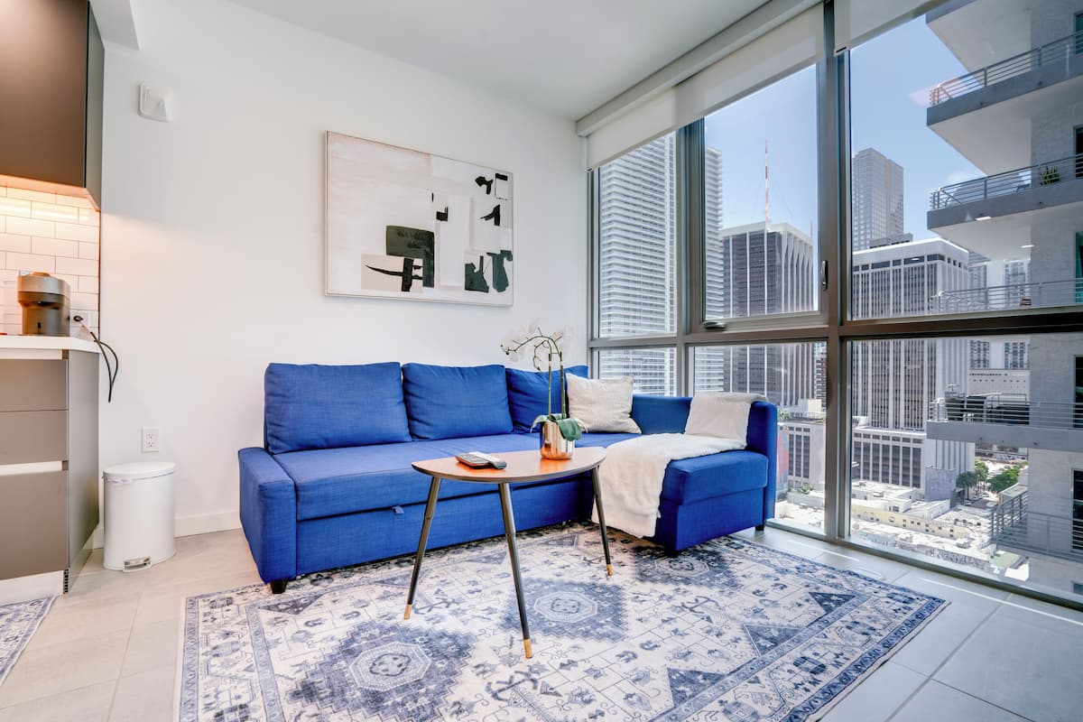 Una sala de estar moderna con un sofá azul, una cafetera, una mesita y un ventanal con vistas a la ciudad.