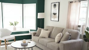 A living room with a beige sofa, a round coffee table, and a white armchair. There is a window with a view of greenery outside. - Cozy Cottage 1.5 mile to  Stadium Ann Arbor (Ann Arbor)