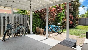 A covered patio area with two bicycles parked under a white pergola, surrounded by lush greenery and a wooden fence.