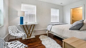 A bedroom with a bed, two windows with blinds, a desk with a lamp, and a chair.