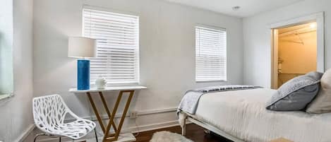 Un dormitorio con cama, dos ventanas con persianas, un escritorio con lámpara y una silla.