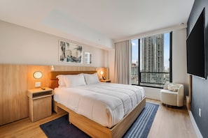 A modern hotel room with a large bed, wooden headboard, bedside tables with lamps, a flat-screen TV, and a large window offering a city view.