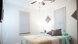 Une chambre à coucher avec un lit, un ventilateur de plafond et deux fenêtres équipées de stores.