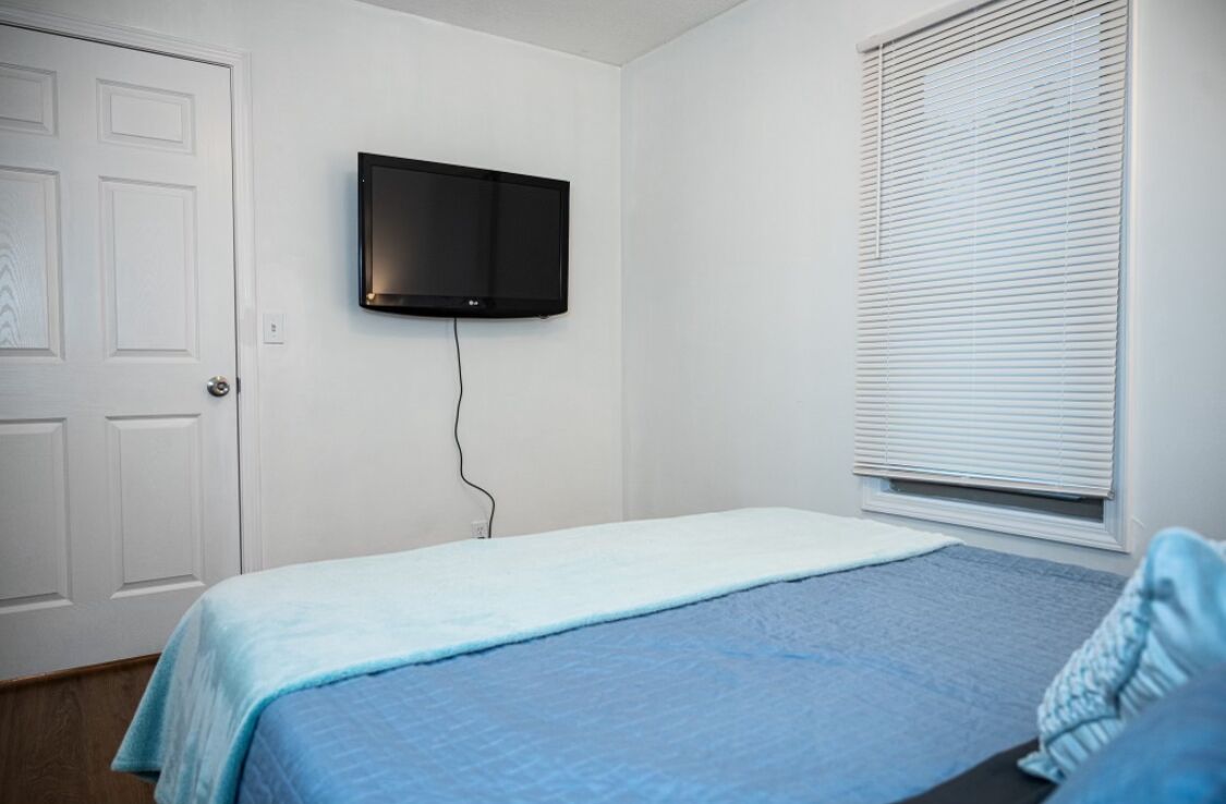 A bedroom with a bed, a wall-mounted TV, a door, and a window with blinds.