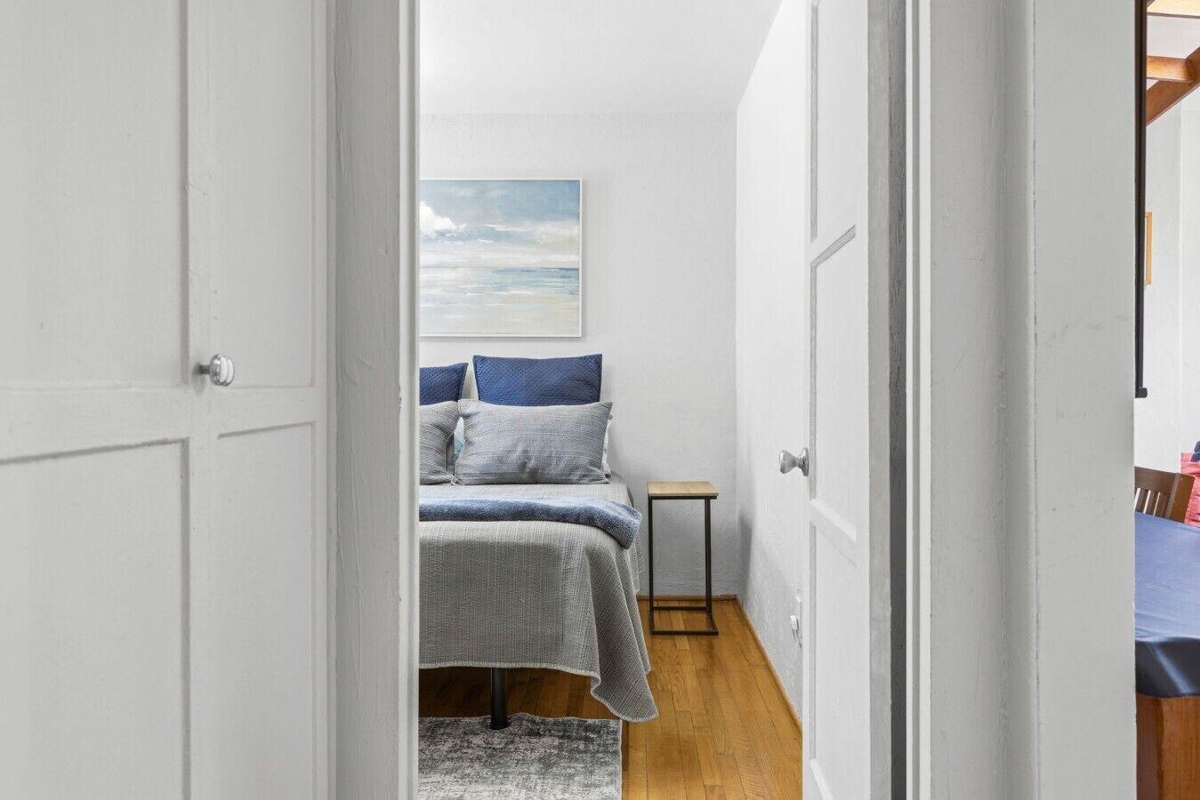 Un couloir étroit mène à une chambre à coucher qui contient un lit, des oreillers, une petite table et un tableau accroché au mur.