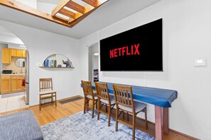 Een moderne woonkamer met een aan de muur bevestigde televisie, een blauwe bank, houten stoelen en een keuken die zichtbaar is door een boog.