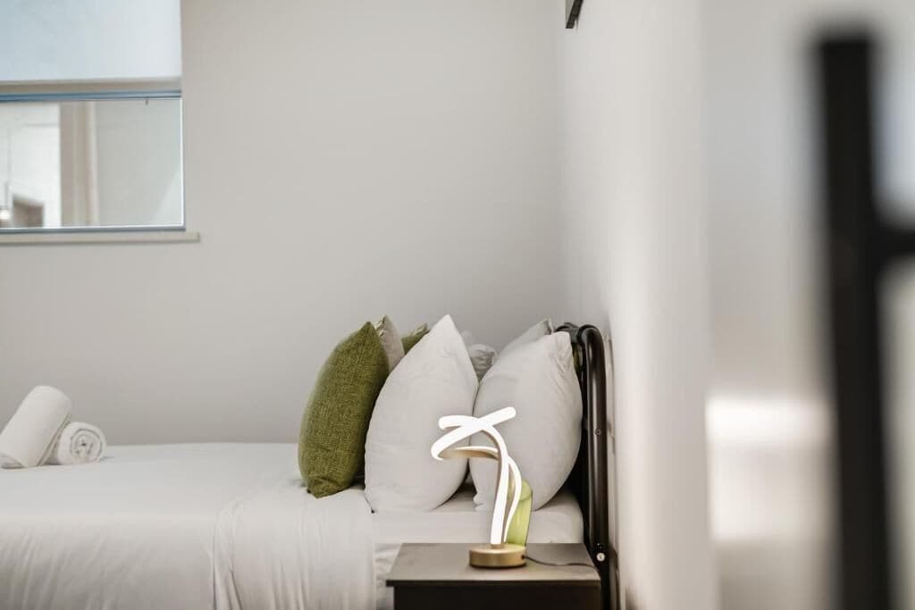 Una cama bien hecha con ropa de cama blanca, una mesita de noche con lámpara y una ventana con persianas.
