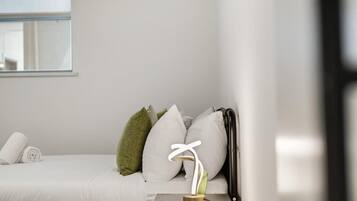 Una cama bien hecha con ropa de cama blanca, una mesita de noche con lámpara y una ventana con persianas.