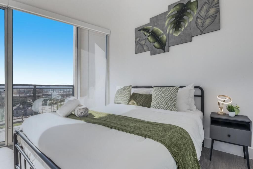 A modern bedroom with a large bed, a nightstand with a lamp, and a view of the city through a large window.