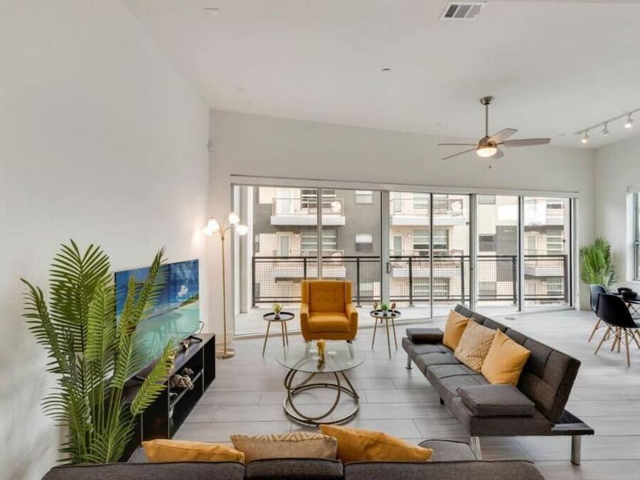 A spacious living area with a sofa, armchair, and coffee table, large windows with a view of buildings, and a balcony with a dining area.