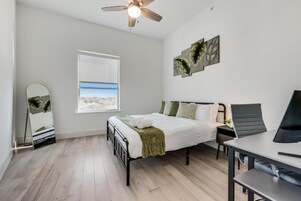 A bedroom with a bed, a desk, a chair, a ceiling fan, and a window with a view.