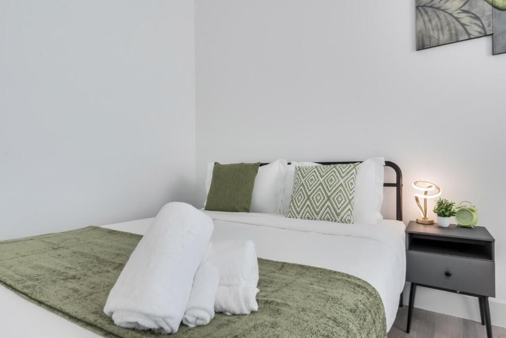 Un lit bien fait, avec du linge de lit blanc et un plaid vert, une table de chevet avec une lampe et une petite plante, et une œuvre d’art accrochée au mur.