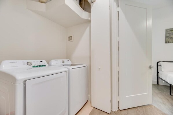 A laundry room with two white Samsung washing machines, a white door, and a bed with white bedding.