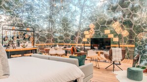 A modern bedroom with a large bed, a desk, and a TV. The room has a wooden floor and a large window with a view of trees. - 1 of a Kind! Largest Luxurious TRANSPARENT DOME in USA (Mars Hill)