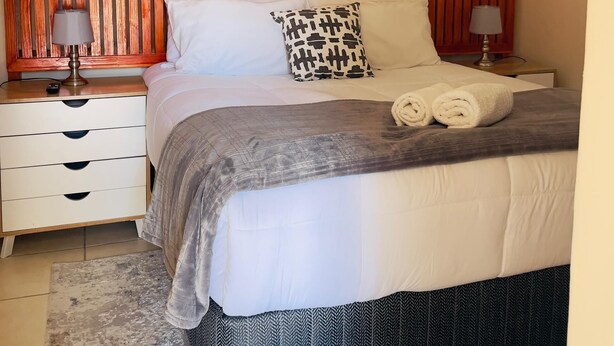 Een netjes opgemaakt bed met een bedspreide met patroon, twee kussens en opgerolde handdoeken op het hoofdeinde.