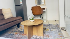 Een moderne woonkamer met een bank, een ronde houten salontafel, een kleine potplant, een koelkast, een stoel en een spiegel.