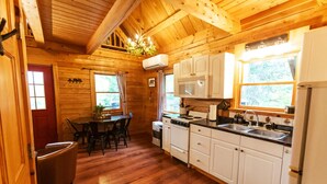 A wooden cabin interior with a dining area, kitchen, and a window with curtains. - Log Cabin Near Bangor and Acadia National Park (Winterport)