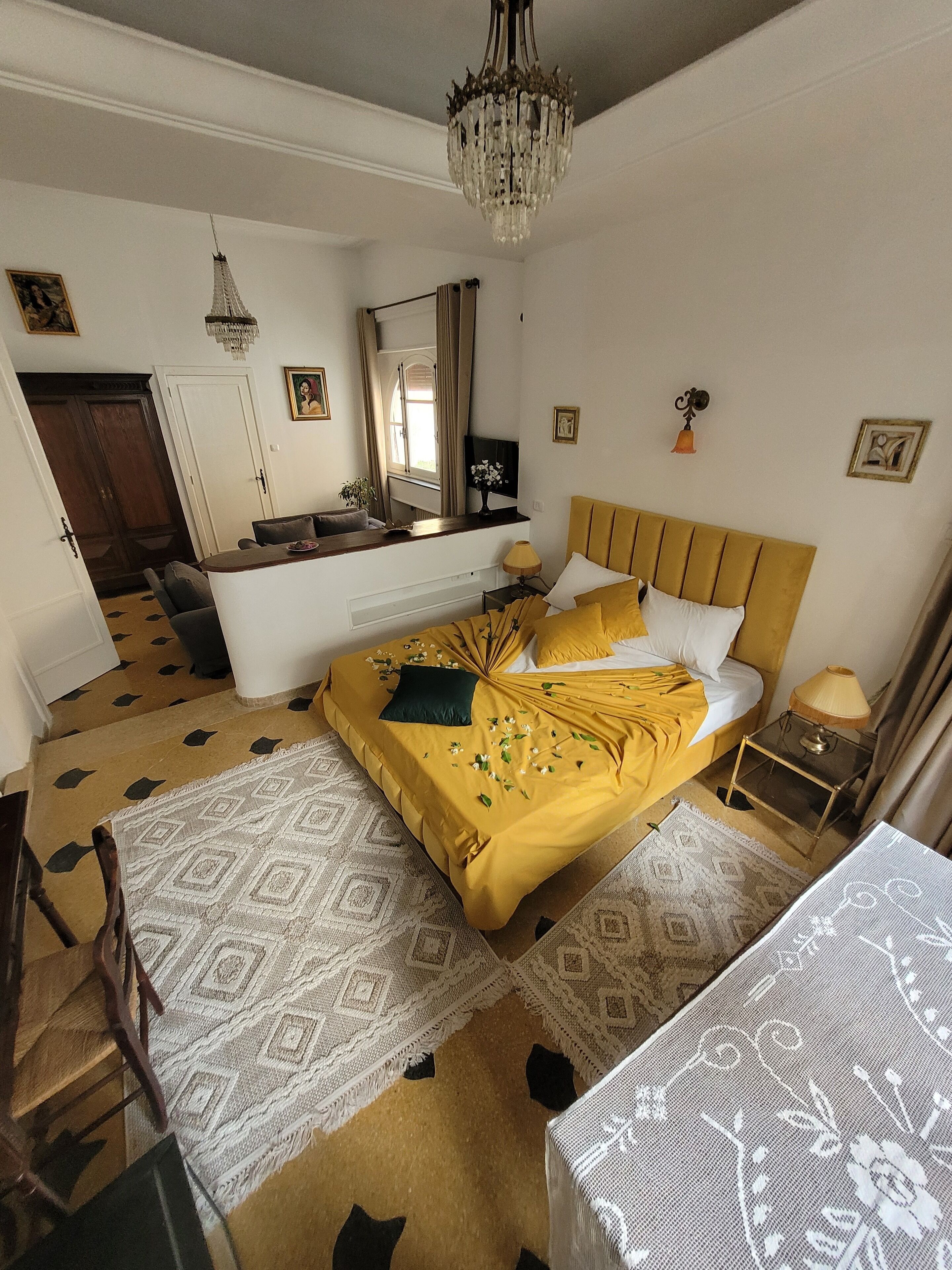 A hotel room with a bed, a desk, a chair, a small table, a lamp, a mirror, and a chandelier.