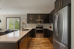 Une cuisine moderne dotée d’un îlot central, d’appareils électroménagers en acier inoxydable et de meubles de rangement aux couleurs sombres.