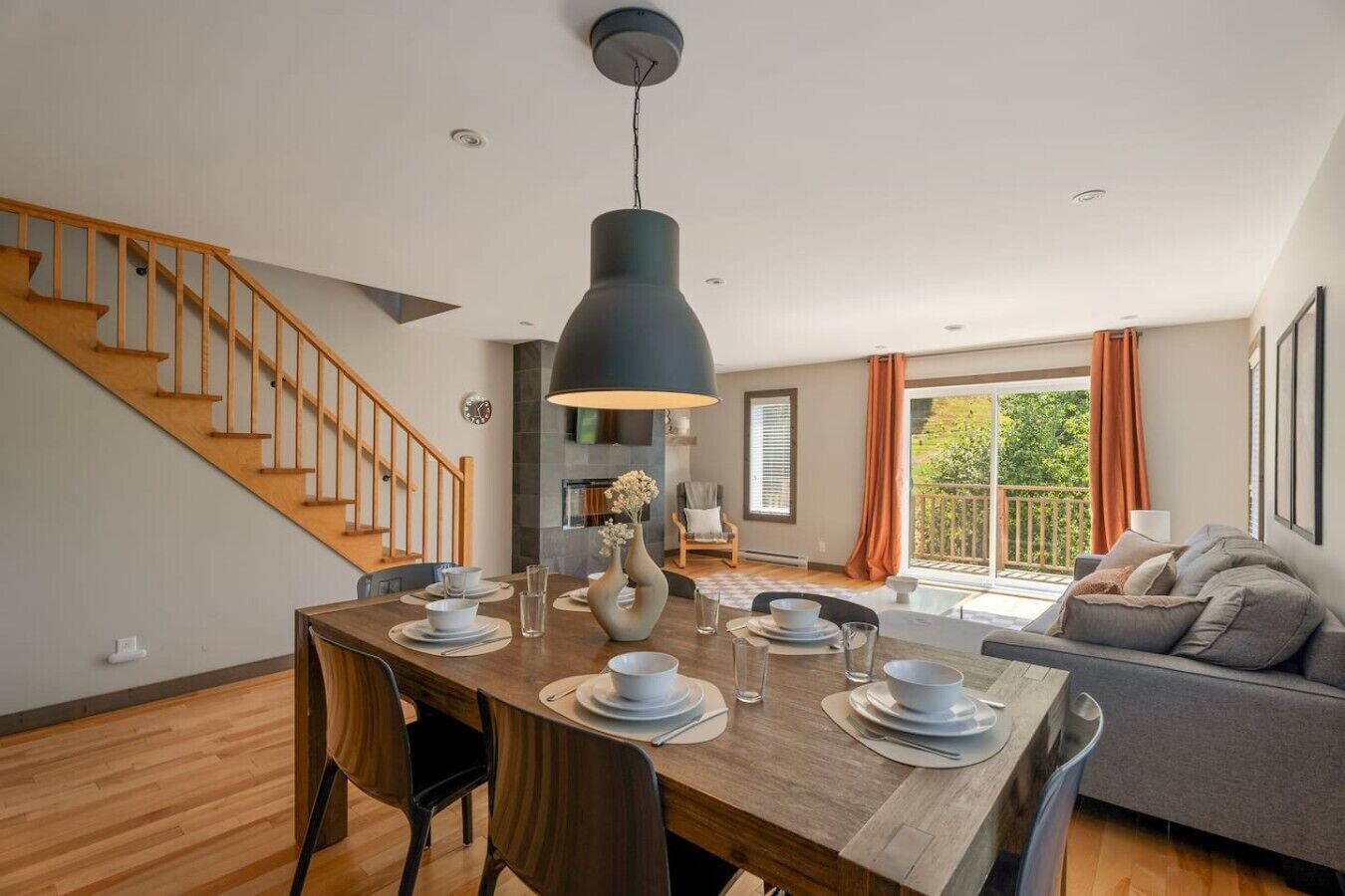 Comedor con una mesa de madera dispuesta para cuatro, una lámpara colgante moderna y una escalera que sube a otro nivel.