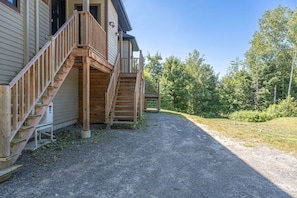 Un bâtiment doté d’un escalier en bois menant à un porche, d’une allée gravillonnée et d’une zone boisée en arrière-plan.