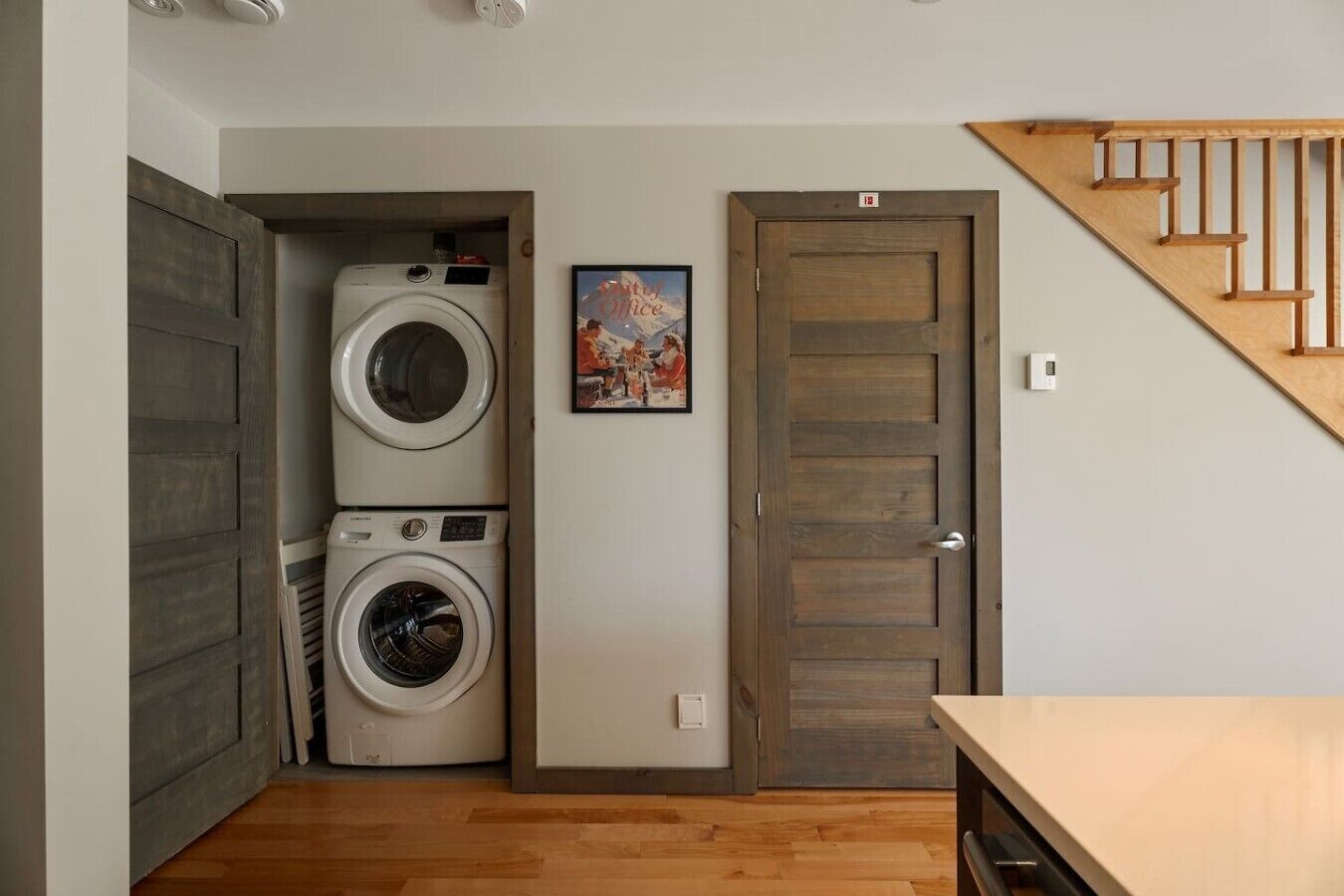 A laundry room with stacked washers and dryers, a wooden door, and a staircase.