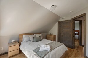 Een slaapkamer met een schuine plafond, een bed met kussens, een nachtkastje, een houten dressoir en een deur naar een andere kamer.